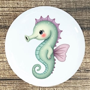 May include: A round, white plate featuring a cartoon seahorse illustration. The seahorse is light green with a pink fin and dorsal fin. It has a cute, smiling face with rosy cheeks and blue eyes.