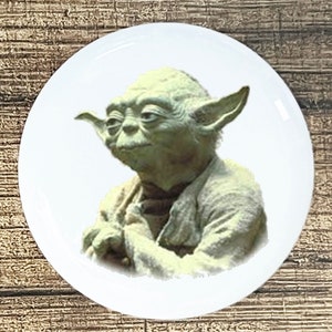 May include: A white ceramic plate featuring a detailed image of Yoda from Star Wars. The image shows Yoda's face and upper body in shades of green and tan. The plate has a glossy finish and a round shape.