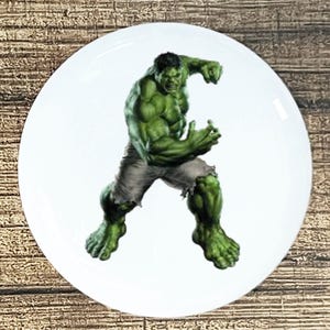 May include: A round white ceramic plate featuring a central graphic illustration of the Hulk, a muscular green humanoid character with short dark hair, wearing tattered grey shorts and posed in a dynamic, aggressive stance with clenched fists.
