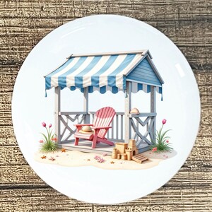 May include: A decorative plate featuring a beach scene. A pink Adirondack chair sits under a blue and white striped awning. The scene includes sand, a sandcastle, seashells, and a straw hat. The plate has a white border and is set against a wood grain background.
