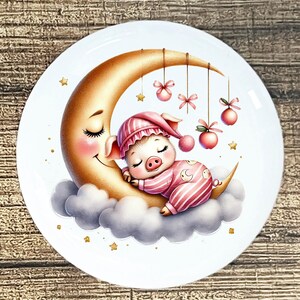May include: A white ceramic plate featuring a whimsical illustration of a sleeping piglet in pink striped pajamas and a matching cap, nestled on a crescent moon. The moon has a smiling face and is surrounded by clouds. Decorative elements include hanging pink bows, stars, and apples.