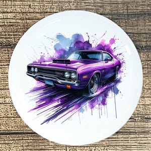 May include: A white ceramic plate featuring a purple muscle car illustration. The car is depicted in a dynamic, artistic style with purple and blue watercolor-like splashes. The car has a black hood scoop and chrome accents.