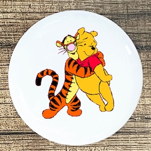 May include: A white button with a cartoon image of Winnie the Pooh hugging Tigger. Tigger is orange and black with a red nose. Winnie the Pooh is yellow with a red shirt.