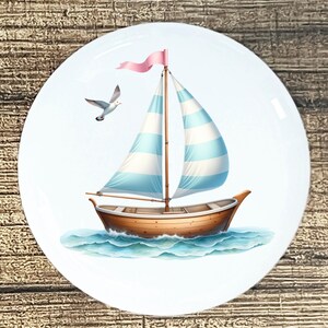 May include: A white ceramic plate featuring a nautical design. A brown sailboat with blue and white striped sails floats on blue water. A seagull flies nearby, and a pink pennant tops the mast.