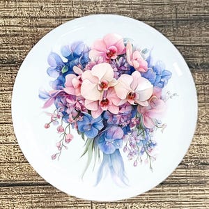May include: A white ceramic plate featuring a watercolor floral design. The design showcases a bouquet of pink and white orchids, blue hydrangeas, and purple flowers. The plate is round and sits on a wooden surface.