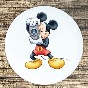 May include: A white ceramic plate featuring a cartoon illustration of Mickey Mouse holding a gray camera. Mickey Mouse is wearing red shorts, yellow shoes, white gloves, and a black top. The plate has a glossy finish and a round shape.
