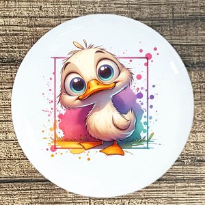 May include: A white ceramic plate featuring a cartoon duckling with large blue eyes and an orange beak. The duckling is set against a colorful, abstract background with a pink and purple square. The plate has a glossy finish.