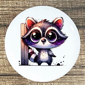 May include: A white ceramic plate featuring a cartoon raccoon with large purple eyes and a black and white striped tail. The raccoon is leaning against a wooden post. The design has a whimsical, illustrated style.