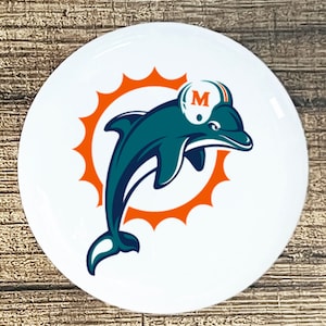May include: A white round button with a teal dolphin jumping out of an orange sunburst. The dolphin is wearing a white helmet with a blue "M" on it.
