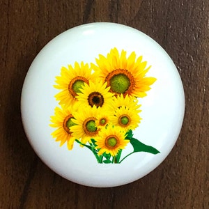 May include: A white ceramic knob with a design of a bouquet of yellow sunflowers with green leaves.
