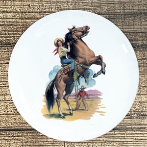 May include: White ceramic plate featuring a vintage illustration of a cowboy riding a rearing brown horse. The cowboy wears a hat, red neckerchief, and blue jeans. A second cowboy stands in the background. The plate has a glossy finish.