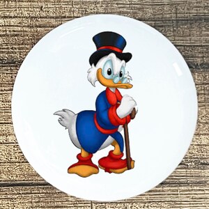 May include: White ceramic plate featuring a cartoon illustration of Scrooge McDuck. The character is depicted in a blue coat, red bow tie, and black top hat, holding a cane. The plate has a glossy finish.