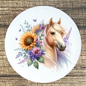 May include: A white round decorative plate featuring a horse's head with a flowing mane. The horse is surrounded by sunflowers, purple flowers, and green leaves. Butterflies are scattered around the design. The plate is set against a wooden background.
