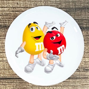 May include: A white round sticker with a yellow and red M&M's character. The yellow M&M's character is wearing white gloves and is holding a red M&M's character. Both characters have a white "M" on their bodies.