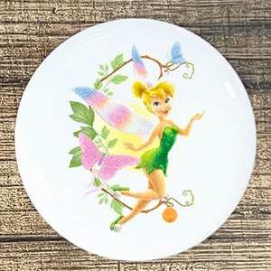 May include: White ceramic plate featuring a colorful illustration of Tinkerbell, a fairy with blonde hair and green dress. The design includes butterflies, leaves, and a branch. The plate has a smooth, glossy finish.