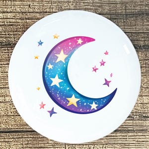 May include: White ceramic plate featuring a crescent moon design with a gradient of pink, blue, and purple. The moon is adorned with yellow and white stars of varying sizes. Additional pink and purple stars are scattered around the moon.
