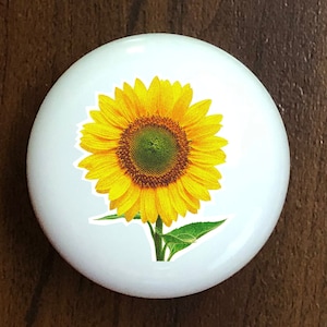 May include: A white ceramic tile with a yellow sunflower with green leaves printed on it.
