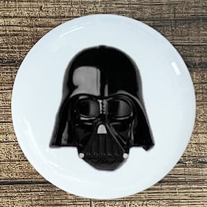 May include: A white round button with a black and white image of Darth Vader's helmet.
