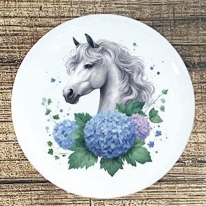 May include: White ceramic plate featuring a watercolor illustration of a white horse's head with a flowing mane. The horse is surrounded by blue and purple hydrangeas and green foliage. Decorative plate.