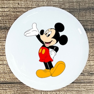 May include: A white ceramic button with a cartoon image of Mickey Mouse. Mickey is wearing his signature red shorts, yellow shoes, and white gloves. He is smiling and waving with his right hand.