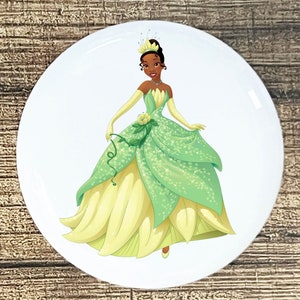 May include: A round sticker featuring a cartoon illustration of Princess Tiana from Disney's The Princess and the Frog. She is wearing a green and yellow ball gown and a crown.