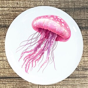 May include: A round, white decorative plate featuring a vibrant pink jellyfish illustration. The jellyfish has a bell-shaped body with white spots and long, flowing tentacles in shades of pink and purple. The plate is set against a wooden background.