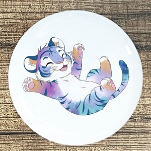 May include: A white, round ceramic plate featuring a cartoon tiger cub. The tiger has a galaxy-themed design with blue, purple, and pink stripes. The cub is lying on its back, smiling, with its paws in the air.