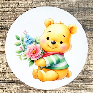 May include: A white ceramic plate featuring a cartoon bear holding a bouquet of flowers. The bear is yellow with a green and white striped shirt and a red scarf. The flowers are pink, blue, and green.