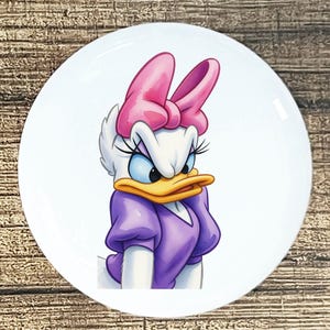 May include: White ceramic plate featuring a cartoon illustration of Daisy Duck. Daisy is depicted with a pink bow, purple dress, and a stern expression. The plate has a glossy finish and is set against a wooden background.