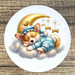 May include: A white ceramic plate featuring a sleeping corgi puppy wearing a blue pajama set with cloud patterns and a matching cap. The puppy rests on a cloud in front of a golden crescent moon. Decorative gold-colored bow-shaped ornaments hang from above.