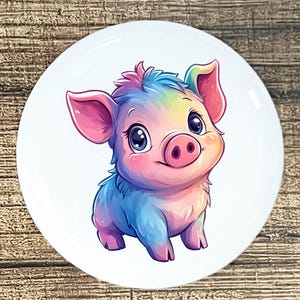 May include: A round, white ceramic plate featuring a colorful illustration of a cartoon pig. The pig has a rainbow-colored coat, pink ears, and a pink snout. The pig is smiling and has large, blue eyes.