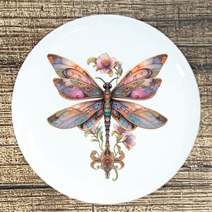 May include: A white ceramic plate featuring a detailed illustration of a dragonfly with colorful wings in shades of orange, pink, blue, and purple. The dragonfly is surrounded by pink flowers and decorative elements.