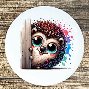 May include: A white ceramic plate featuring a cartoon hedgehog with large blue eyes, colorful quills, and a friendly expression. The hedgehog peeks from behind a wooden post, with colorful paint splatters on the right side.