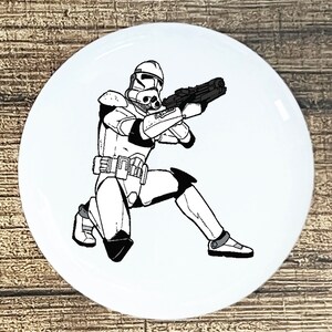 May include: A black and white illustration of a Star Wars clone trooper kneeling and aiming a blaster rifle. The trooper is wearing a helmet and armor.