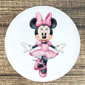 May include: A white ceramic plate featuring a cartoon illustration of Minnie Mouse in a pink ballerina outfit. Minnie Mouse is wearing a pink dress, ballet shoes, and a bow. The plate has a glossy finish and a round shape.