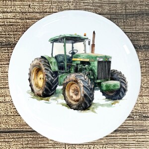 May include: A white ceramic plate featuring a watercolor painting of a green tractor with large tires. The tractor has a yellow stripe and is set against a grassy background. The plate is round and has a glossy finish.