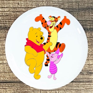 May include: A white round button with a cartoon image of Winnie the Pooh, Tigger, and Piglet. Winnie the Pooh is wearing a red shirt and is yellow. Tigger is orange and black with a yellow belly. Piglet is pink with a striped shirt.