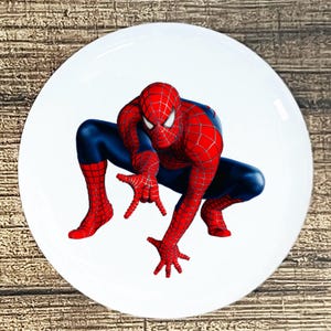 May include: White ceramic plate featuring a vibrant image of Spider-Man in his iconic red and blue suit. The superhero is depicted in a crouching pose, with his hands outstretched. The plate has a glossy finish.