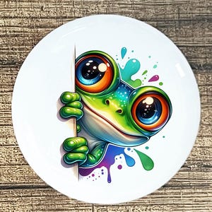 May include: A white ceramic plate featuring a cartoon frog with large, colorful eyes. The frog is green, blue, and orange, peeking from behind a white object. Splashes of color surround the frog, adding to the playful design. The plate is on a wooden surface.