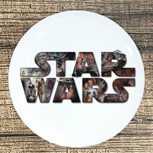 May include: A white, round plate featuring the words "STAR WARS" in a bold, stylized font. The letters are filled with images of characters and scenes from the Star Wars films. The plate has a glossy finish.