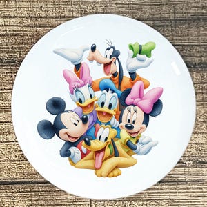 May include: White ceramic plate featuring a colorful illustration of Disney characters Mickey Mouse, Minnie Mouse, Donald Duck, Daisy Duck, Goofy, and Pluto. The plate has a glossy finish and is suitable for serving food.