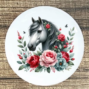 May include: A white ceramic plate featuring a detailed illustration of a horse's head with a flowing mane. The horse is surrounded by red, pink, and blue flowers, with green leaves and small butterflies. The design is centered on the plate.