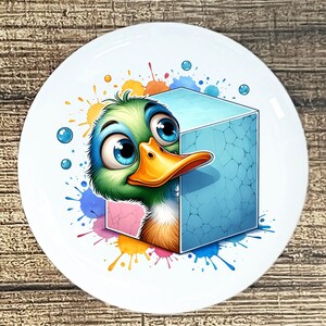 May include: A white ceramic plate featuring a cartoon duck with large blue eyes and an orange beak. The duck is peeking out from behind a blue cube. Colorful paint splatters and bubbles surround the duck and cube.