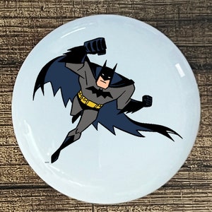 May include: A white round button with a cartoon image of Batman in a flying pose. Batman is wearing a black cape and suit with a yellow belt. The background is light blue.