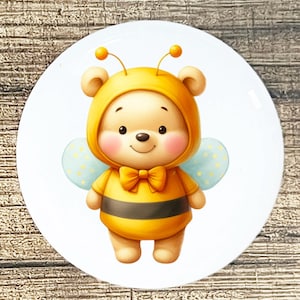 May include: A white ceramic plate featuring a cartoon bear dressed as a bee. The bear wears a yellow hooded outfit with a black stripe, a bow tie, and light blue wings with yellow polka dots. The bear has rosy cheeks and a smiling expression.
