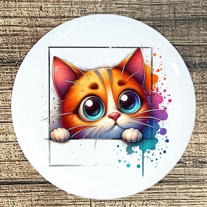 May include: A white ceramic plate featuring a cartoon cat with orange and white fur, large blue eyes, and pink ears. The cat is framed by a black square, with colorful paint splatters in the background. The plate has a glossy finish.