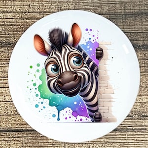 May include: A white ceramic plate featuring a cartoon zebra with black and white stripes, large blue eyes, and a friendly expression. The zebra peeks from behind a brick wall, with colorful paint splatters in the background.
