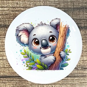 May include: A white ceramic plate featuring a cartoon koala bear. The koala is gray with large brown eyes, holding onto a tree branch. Green leaves and colorful paint splatters surround the koala, creating a whimsical design.