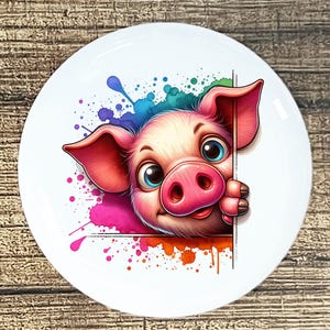 May include: White ceramic plate featuring a cartoon pig with a pink snout, ears, and big blue eyes. The pig is peeking out from behind a white border, with colorful paint splatters in pink, orange, blue, and purple.