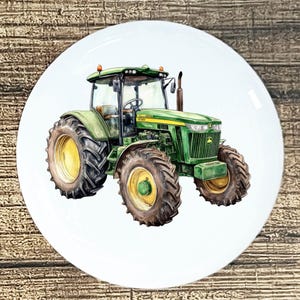 May include: A white ceramic plate featuring a watercolor illustration of a green tractor with yellow wheels. The tractor has a detailed design, including headlights, a cab, and a smokestack. The plate is round and has a glossy finish.
